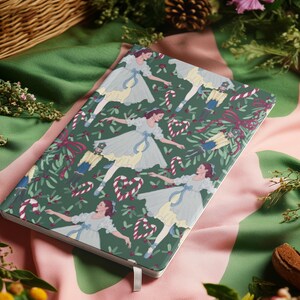 Pode incluir: Um caderno de capa dura com uma capa verde com um padrão de bailarinas, bengalas doces e quebra-nozes. O caderno tem uma fita branca como marcador e está sobre um tecido rosa e verde.