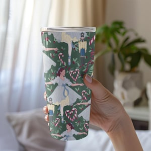 May include: A tall, insulated tumbler with a festive Nutcracker-themed design. The tumbler features illustrations of ballerinas, toy soldiers, candy canes, and heart-shaped candies on a dark green background. The design is repeated around the entire tumbler.