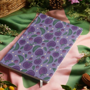 Puede incluir: Un cuaderno rectangular con una cubierta morada que presenta un patrón de ciruelas moradas, hojas verdes y pequeñas flores blancas. El cuaderno tiene una cinta blanca como marcador y está sobre una tela rosa y verde.