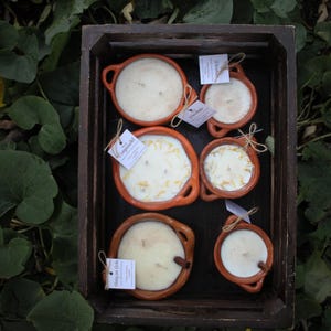 May include: A wooden crate contains several terracotta candles with white wax. Some candles have dried flower petals. Each candle has a small tag attached with twine. The candles are various sizes and shapes.