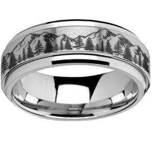 May include: A silver-colored ring with a mountain and forest design. The ring features a detailed engraving of a mountain range and trees around the band. The ring has a polished, reflective surface.
