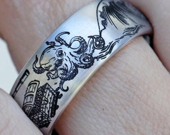 May include: A silver ring with a black and white engraved design of an octopus and a wave. The ring is worn on a person's finger.
