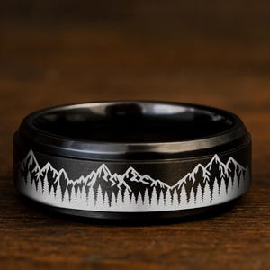 Peut inclure: Une bague noire avec un motif de montagne et de forêt blanc. La bague est en métal foncé et repose sur une surface en bois. Le design de la bague présente une chaîne de montagnes et des arbres détaillés.