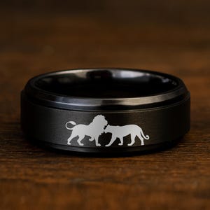 May include: A black ring with a matte finish. The ring features a design of two lions in a light color. The ring is set against a dark wooden surface.