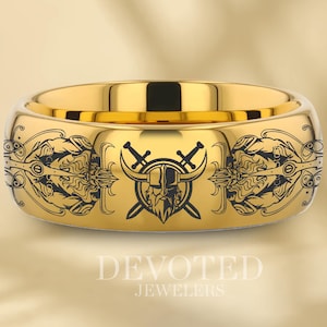 Może przedstawiać: Złoty pierścionek z czarnym grawerowanym wzorem przedstawiającym głowę z rogami, mieczami i ozdobnym wzorem. Pierścionek jest grawerowany tekstem "DEVORED JEWELERS".