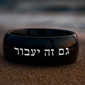 May include: A black ring with the Hebrew text "Gam Zeh Ya'avor" in white lettering. The ring is resting on sand, with a blurred ocean background.