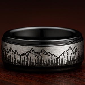 Peut inclure: Une bague noire et argentée avec un motif de montagne et de forêt. La bague présente un anneau noir et un anneau argenté avec une ligne de montagnes et d'arbres détaillée gravée autour de la bague. La bague est sur une surface en bois.