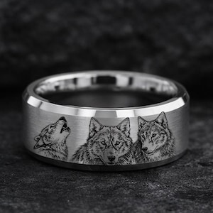 May include: A silver-colored ring with a detailed engraving of three wolves. The ring features a flat, wide band with the wolf images encircling the exterior. One wolf is howling, and the other two are facing forward.
