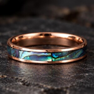 May include: A rose gold band ring featuring a central inlay of iridescent abalone shell. The ring has a polished metallic finish with a smooth, rounded profile, showcasing the natural blue and green color variations of the shell segments.