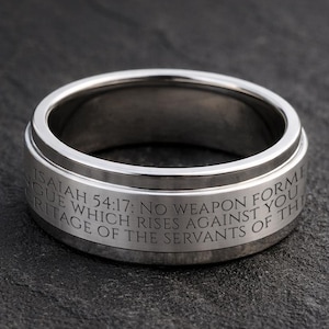 May include: A silver-colored ring with the engraved text "ISAIAH 54:17: NO WEAPON FORMED AGAINST YOU WHICH RISES AGAINST YOU HERITAGE OF THE SERVANTS OF THIS". The ring is circular and has a polished finish.