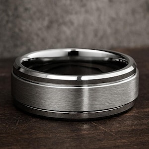 May include: A silver-colored ring with a brushed metal center band and polished edges. The ring is set against a dark wooden surface, showcasing its metallic sheen and detailed design. This ring is a piece of jewelry.