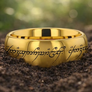 Op de afbeelding: Een goudkleurige ring met zwarte gegraveerde tekst. De ring ligt op donkere grond, met een vage groene achtergrond. Het ontwerp van de ring is een eenvoudige band.