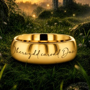 May include: A polished gold band ring featuring a smooth, reflective surface and a rounded profile. The exterior of the ring is engraved with the black cursive script text "Oherwydd carodd Duw" across the front center of the metal surface.