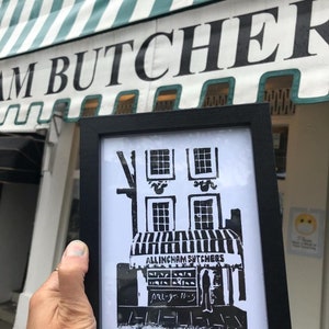 May include: A black and white framed print of a butcher shop with the text "ALLINGHAM BUTCHERS". The print is held in front of a building with the word "BUTCHER" visible on the awning. The print depicts a two-story building with windows and a striped awning.