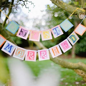 May include: A two-tiered decorative banner featuring square paper cards strung together. The cards display colorful, block-printed letters spelling out the phrase JUST MARRIED, interspersed with patterned squares in shades of pink, blue, and yellow.