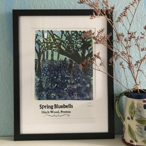 Op de afbeelding: Een ingelijste print van een blauwe bos scène met de titel "Spring Bluebells Hitch Wood, Preston" gedrukt onder de afbeelding.