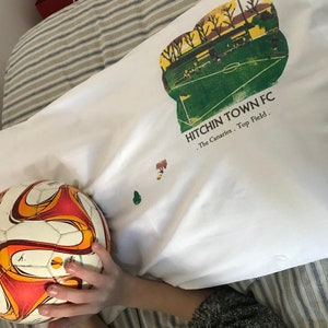 Könnte beinhalten: Ein weißes Kissen mit einer grünen und gelben Grafik eines Fußballfeldes und dem Text "Hitchin Town FC. The Canaries. Top Field." Ein Fußball befindet sich im Vordergrund.
