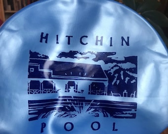 May include: A light blue swimming cap with the text "Hitchin Pool" and a black and white illustration of a building with a pool in front of it.