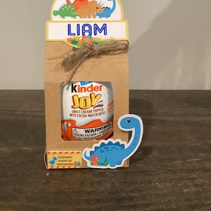Può includere: Una scatola regalo di carta marrone con un uovo Kinder Joy, un adesivo di dinosauro blu e un'etichetta con il nome "LIAM". La scatola è decorata con un fiocco di spago e un topper a tema dinosauri.