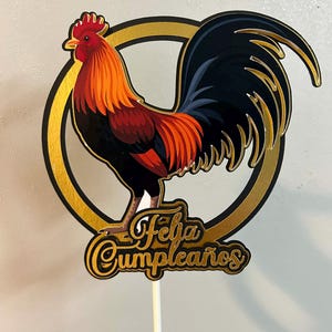 May include: A colorful rooster cake topper with the text "Feliz Cumpleaños". The rooster is depicted in shades of red, orange, and black, set against a gold and black circular frame. The topper is attached to a wooden stick.