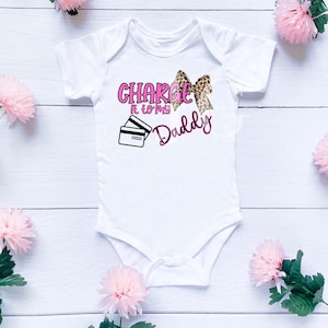 May include: White baby onesie with the text "Charge It To My Daddy" in pink and black. The design includes a leopard print bow and a graphic of two credit cards. Pink flowers are scattered around the onesie.