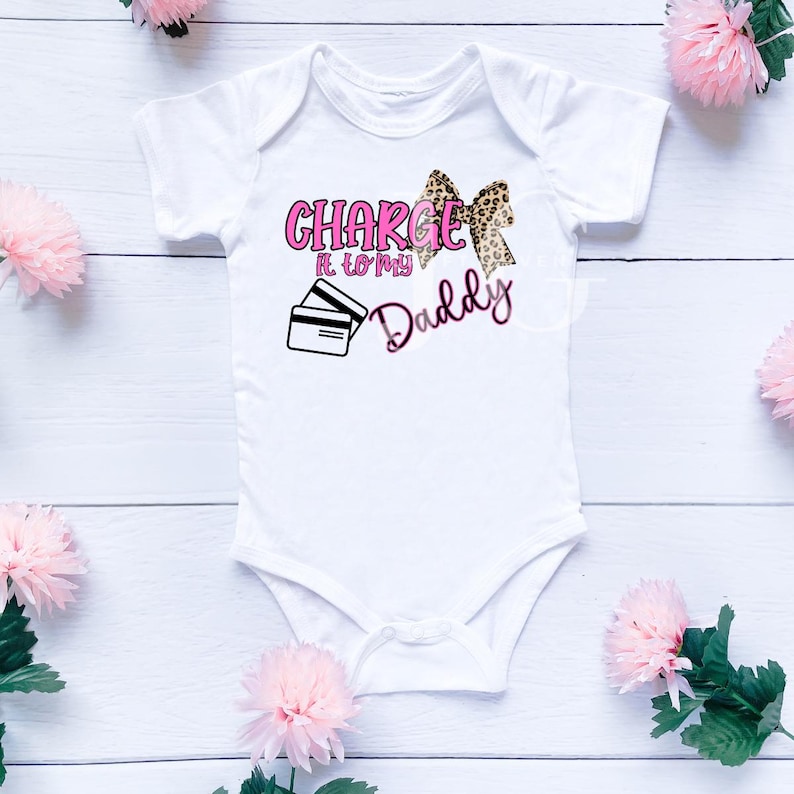 May include: White baby onesie with the text "Charge It To My Daddy" in pink and black. The design includes a leopard print bow and a graphic of two credit cards. Pink flowers are scattered around the onesie.