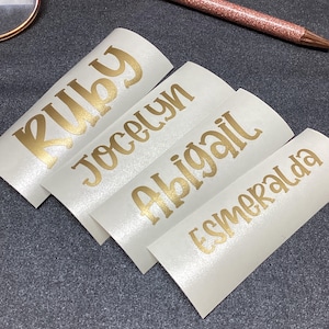 Op de afbeelding: Vier witte zelfklevende labels met metallic gouden letters die de namen Ruby, Jocelyn, Abigail en Esmeralda in een cursief lettertype weergeven, vergezeld door een bril met roségouden montuur en een bijpassende roségouden glitterpen.