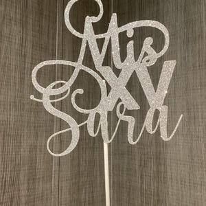 Peut inclure: Un cake topper argenté et scintillant avec l'inscription "Mis XV Sara" en écriture cursive. Le topper est fixé à un bâton blanc et placé sur un arrangement floral rose. Le fond est un grain de bois foncé.