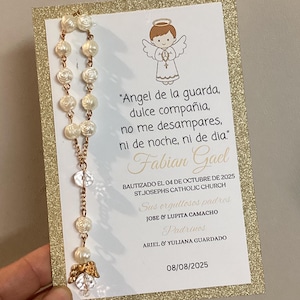 May include: A baptism invitation card with a gold glitter border and a rosary. The card features an illustration of an angel and text in Spanish. The rosary has white rose-shaped beads and gold-coloured accents.