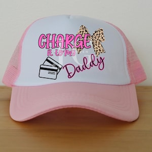 May include: A pink and white trucker hat with the text "CHARGE it to my Daddy" in pink, along with a leopard print bow and credit card graphic. The hat has a pink brim and mesh back.