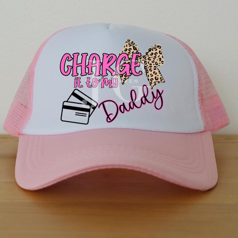 May include: A pink and white trucker hat with the text "CHARGE it to my Daddy" in pink, along with a leopard print bow and credit card graphic. The hat has a pink brim and mesh back.