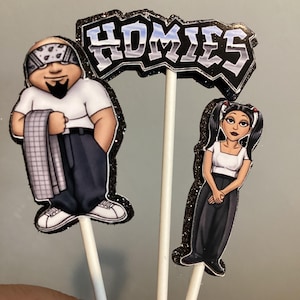 Könnte beinhalten: Kuchenaufsätze mit Cartoonfiguren eines Mannes mit Bandana und einer Frau mit Zöpfen, sowie einem Schild mit der Aufschrift "HOMIES". Die Figuren und das Schild sind mit Glitzer umrandet.