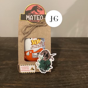 Può includere: Una scatola regalo di carta marrone legata con spago, a tema dinosauri. Include un uovo Kinder Joy, un adesivo di dinosauro e un'etichetta con il nome "MATEO". È presente anche un'etichetta di avvertimento in spagnolo.