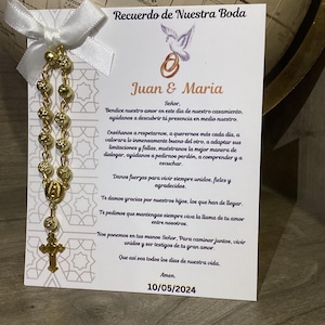 Könnte beinhalten: Ein goldener Rosenkranz mit weißem Band und einer Karte mit der Aufschrift "Recuerdo de Nuestra Boda", den Namen "Juan & Maria" und dem Datum "10/05/2024". Der Rosenkranz hat ein Kreuz und einen kleinen Anhänger.