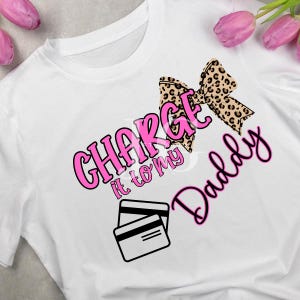 May include: White t-shirt with the phrase "CHARGE it to my Daddy" in pink and black lettering. A leopard print bow and two credit card illustrations are also featured on the shirt. Pink flowers are in the background.