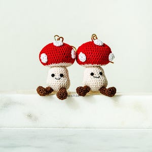 May include: Two crocheted mushroom figures with red caps and white polka dots. The mushroom stems are off-white, with smiling faces and brown feet. Each mushroom has a gold-colored loop at the top.