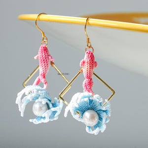 May include: A pair of handmade earrings featuring pink crochet fish suspended from gold-colored hooks. Each earring has a gold square frame with a blue and white crochet wave design and a pearl bead.
