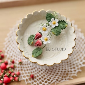 May include: A decorative white ceramic plate with a scalloped gold rim, adorned with a crocheted strawberry and flower design. The plate rests on a white lace doily, with additional crocheted strawberries scattered around. The text "DQ STUDIO" is visible.