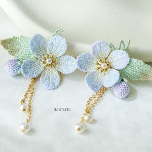 Puede incluir: Un par de pinzas para el pelo con flores hechas a mano. Cada pinza presenta una flor azul claro y blanca con un centro dorado, una hoja verde y una baya morada. Cadenas doradas con gotas de perlas cuelgan de las flores. El texto "DQ STUDIO" es visible.