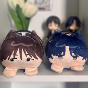 May include: Two plush character pouches with cat ears, one in dark brown and one in dark blue. Each plush features embroidered facial expressions, including eyes and hair details, with a small metal chain attached to the top for hanging purposes.