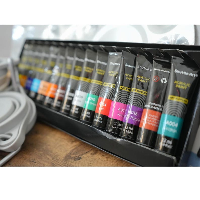 May include: A set of 12 acrylic paint tubes in various colors, including red, purple, green, and blue. The tubes are labeled with the brand name "Shuttle Art" and the color name. Each tube contains 12 ml (0.4 fl oz) of paint.