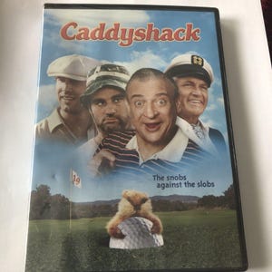 Puede incluir: Carcasa de DVD de la película Caddyshack. La portada presenta un collage de cuatro hombres, un gorgojo en una pelota de golf y el texto "The snobs against the slobs."