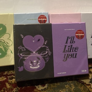 Puede incluir: Una colección de estuches rectangulares de álbumes de música en varios colores, incluyendo amarillo pálido, azul claro, rosa, blanco, negro y morado. Varios estuches presentan el texto I'll Like You y pegatinas rojas circulares exclusivas.