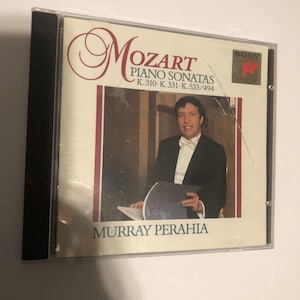 May include: A CD case featuring the album "Mozart Piano Sonatas" by Murray Perahia. The cover has a portrait of Perahia in a tuxedo. The Sony Classical logo is in the upper right corner. The CD case is in a clear plastic case.