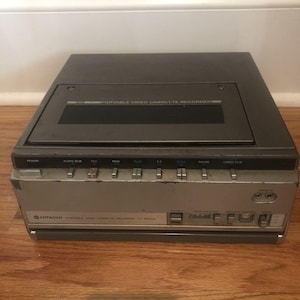 May include: A vintage Hitachi portable video cassette recorder, model VT-9000A, in a silver and dark gray color scheme. The device features various control buttons and a cassette slot. The unit is placed on a wooden surface.