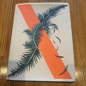 Puede incluir: Una cubierta de libro blanca con una franja naranja diagonal y un diseño de hoja de palmera verde. La hoja de palmera está parcialmente oculta por la franja naranja.