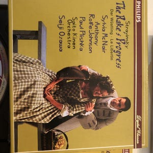 May include: A yellow and brown record album cover with the title "The Rake's Progress" by Stravinsky. The album features a photo of two people in period clothing, a woman in a plaid dress and a man in a brown jacket, with a basket of flowers in the foreground.