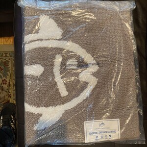 May include: A reversible bath rug in a clear plastic bag. The rug is grey with a white design. The rug has the words "SOUTHERN TIDE" and "REVERSIBLE SKIPJACK BATH RUG" on a white label.