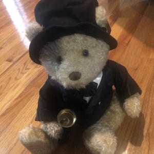 Może przedstawiać: Pluszowy miś w czarnym cylindrze i garniturze, trzymający mały złoty dzwonek. Miś ma brązowe futro i czarną muszkę.