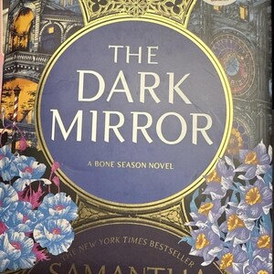 Könnte beinhalten: Ein Hardcover-Buch mit dem Titel "The Dark Mirror" von Samantha Shannon. Das Cover zeigt einen dunkelblauen Kreis mit goldenem Rand, den Titel in Weiß und florale und architektonische Illustrationen. Es ist ein "Bone Season Novel".